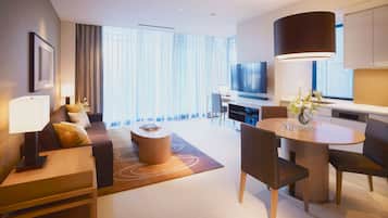 A modern hotel room with a sofa, dining table, chairs, a TV, and a large window with curtains.