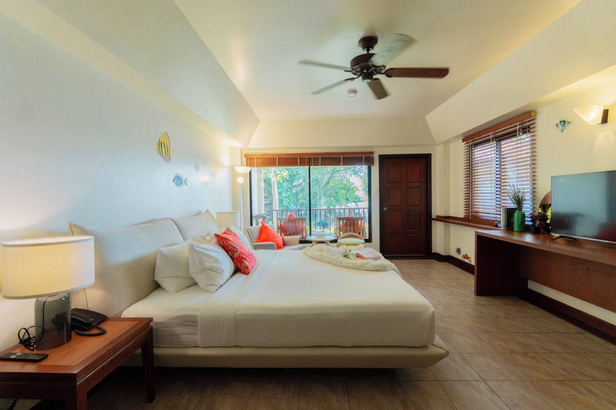 A hotel room with a bed, bedside table, lamp, ceiling fan, TV, and a view of the outdoors.