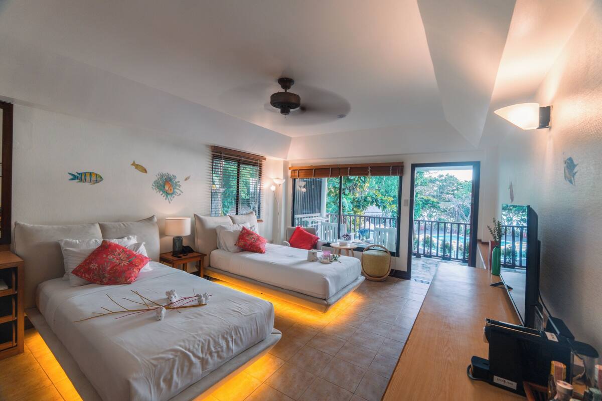 A hotel room with two beds, a TV, a balcony with a view, and a ceiling fan.