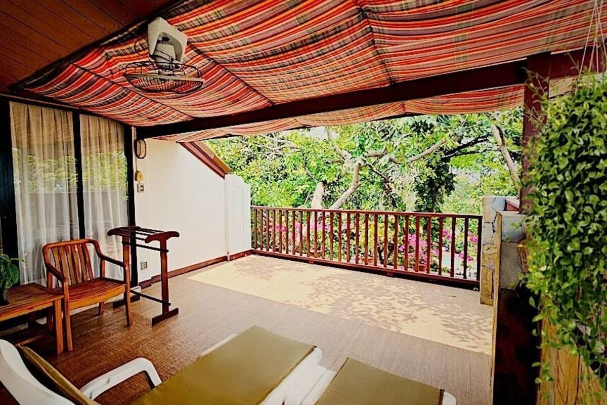 A covered outdoor seating area with wooden furniture, a view of greenery, and a balcony railing.