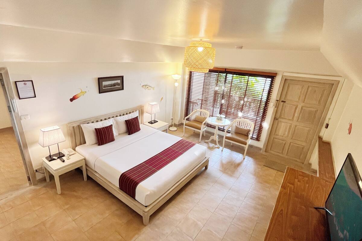 A hotel room with a large bed, a desk with a lamp, a chair, a window with blinds, and a door.