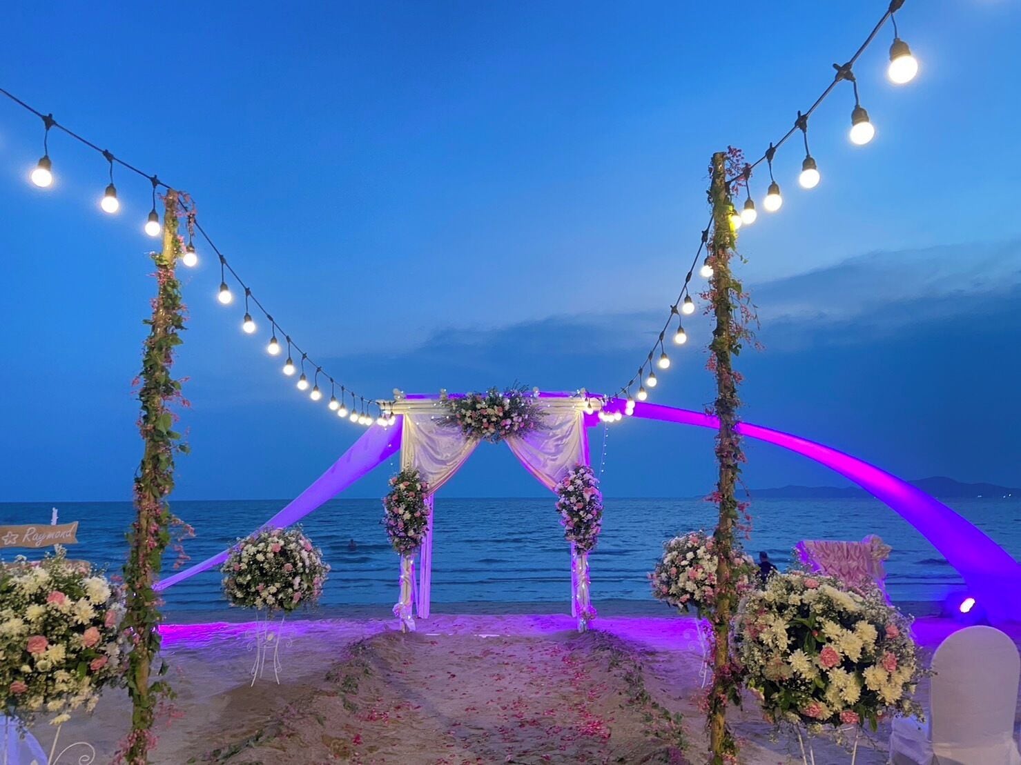 En bröllopsdekoration på stranden med en utsmyckad båge, blomsterarrangemang och ljusslingor.
