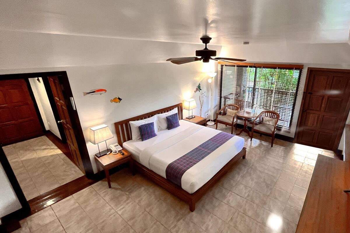 A hotel room with a bed, a desk, a chair, a ceiling fan, and a window with blinds.