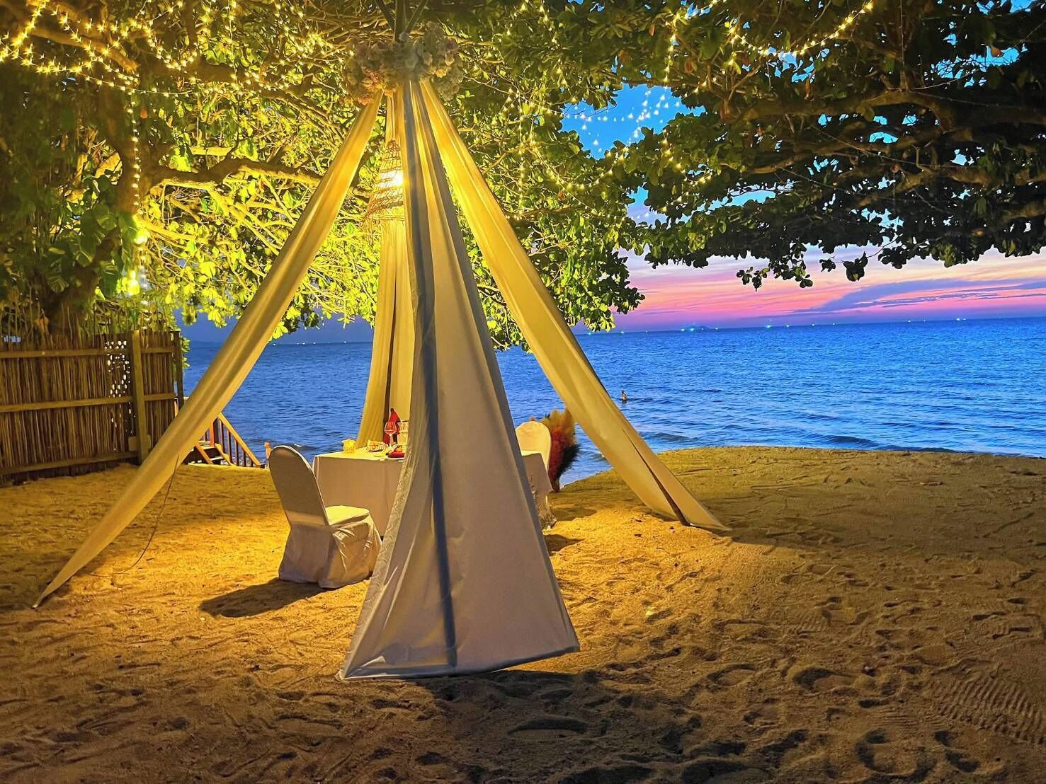 Een strandsetting met een tentachtige constructie, tafels en stoelen onder een boom, met uitzicht op de zee.