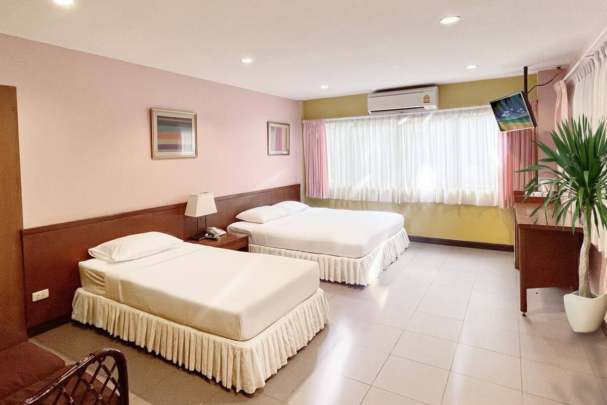 A hotel room with two beds, a desk, a chair, a potted plant, and an air conditioning unit.