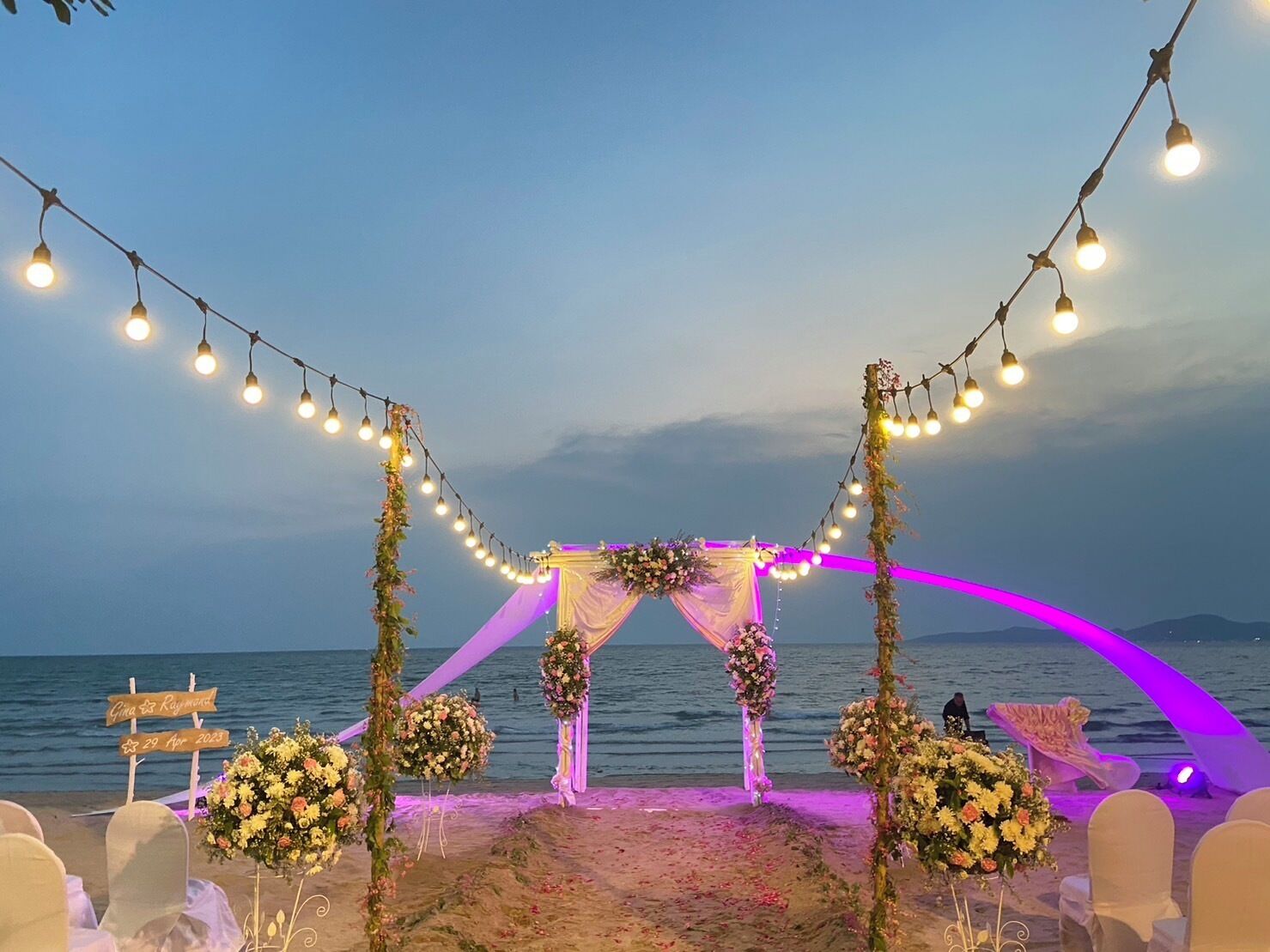 A beach wedding setup with a stage, floral decorations, and string lights.