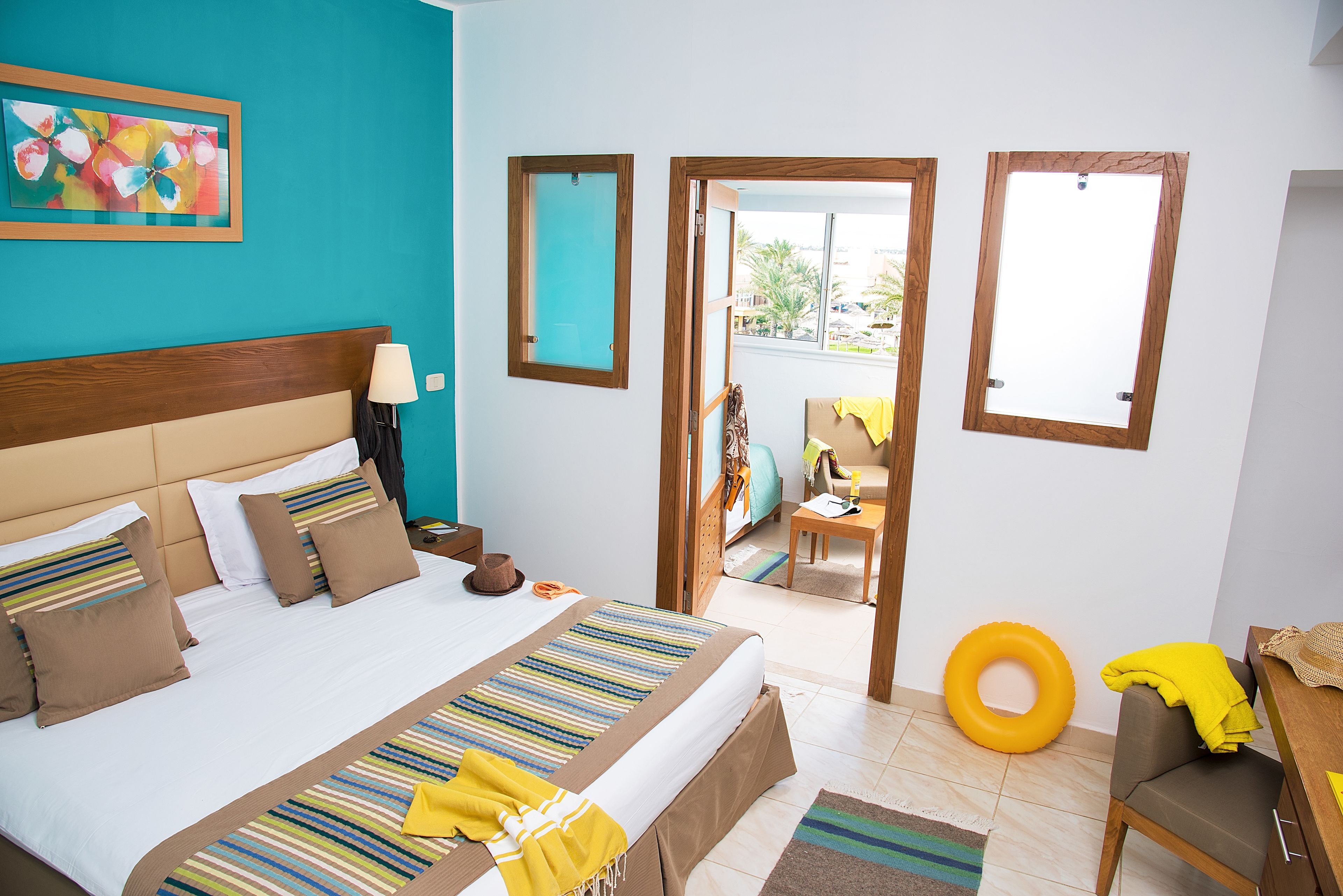 Une chambre d’hôtel avec un grand lit, une tête de lit en bois, une table de chevet avec une lampe, une œuvre d’art colorée au mur et une vue sur le balcon extérieur.