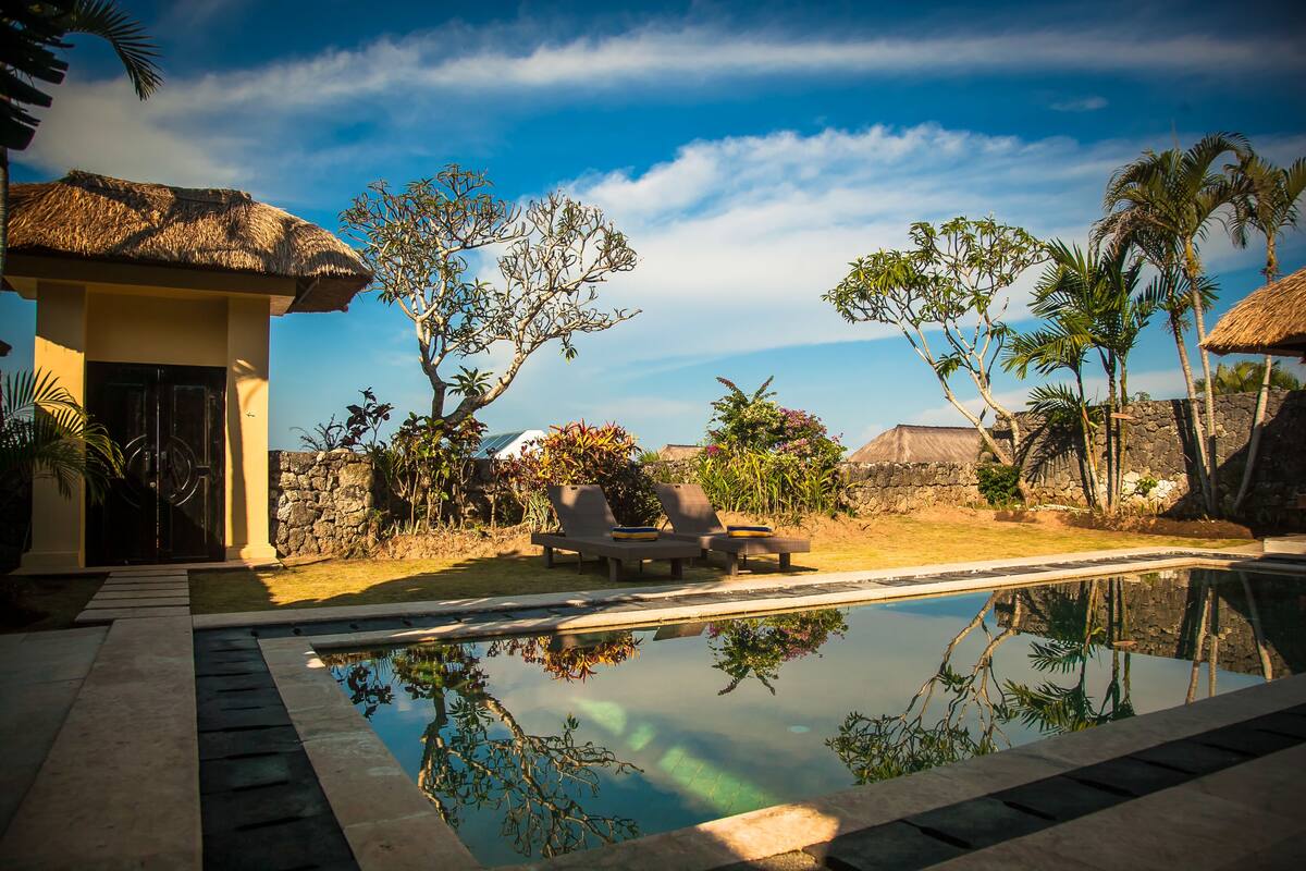 A pool area with lounge chairs, a thatched-roof building, and tropical vegetation.