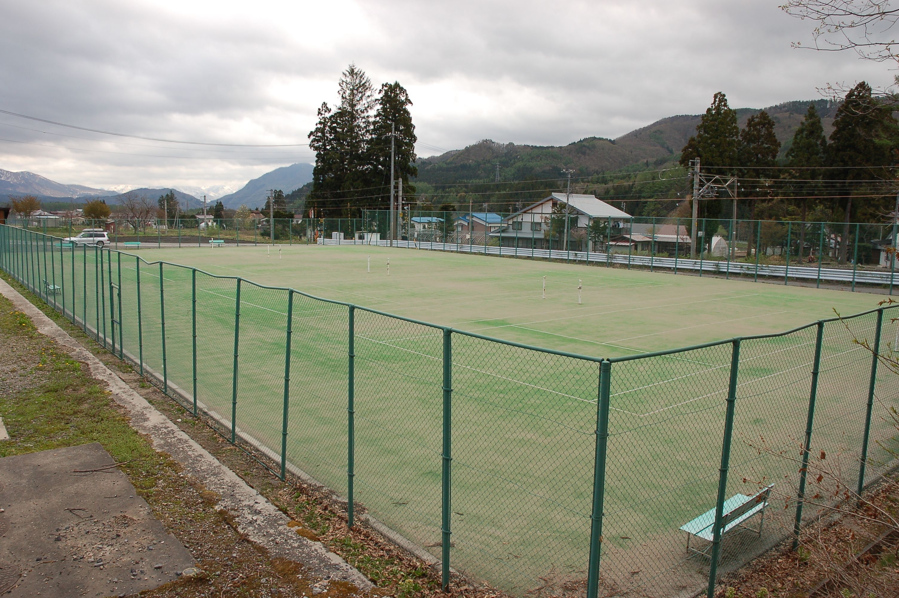Un terrain de sport verdoyant, entouré d’une clôture, avec un banc et des maisons en arrière-plan.