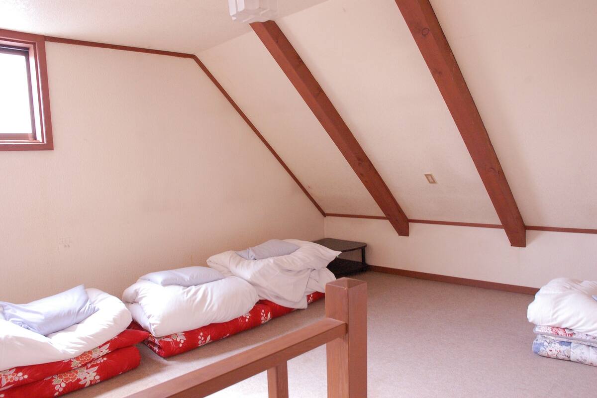Habitació petita individual amb sostre inclinat, una finestra i una cadira de fusta.