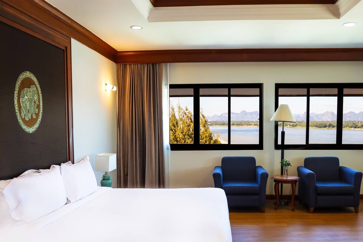 Habitación de hotel con una cama grande, dos sillones, vista a un lago y un panel decorativo en la pared.