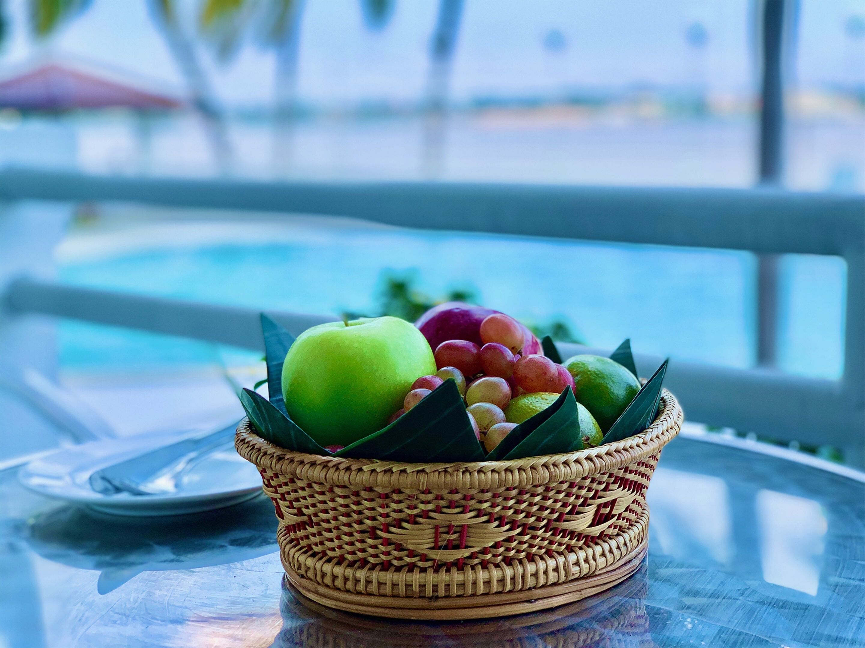 Un panier en osier rempli de fruits est posé sur une table en verre, avec une piscine en arrière-plan.
