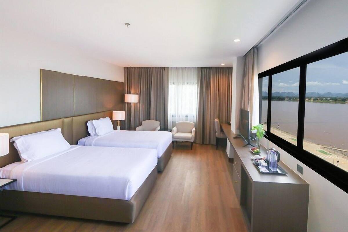 Habitación de hotel con dos camas, un escritorio, una silla, un televisor y un ventanal con vistas al agua.