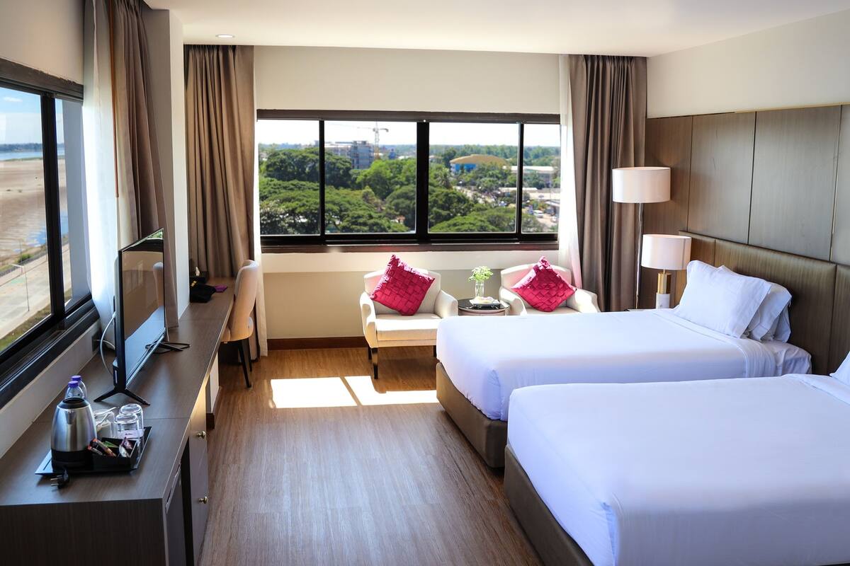 Habitación de hotel con dos camas, un escritorio con televisor de pantalla plana, una ventana con vistas a zonas verdes y edificios, y una lámpara en la mesita de noche.
