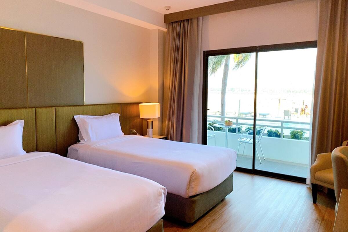 Habitación de hotel con dos camas, un ventanal con vistas a la piscina y palmeras, y una silla junto a la ventana.