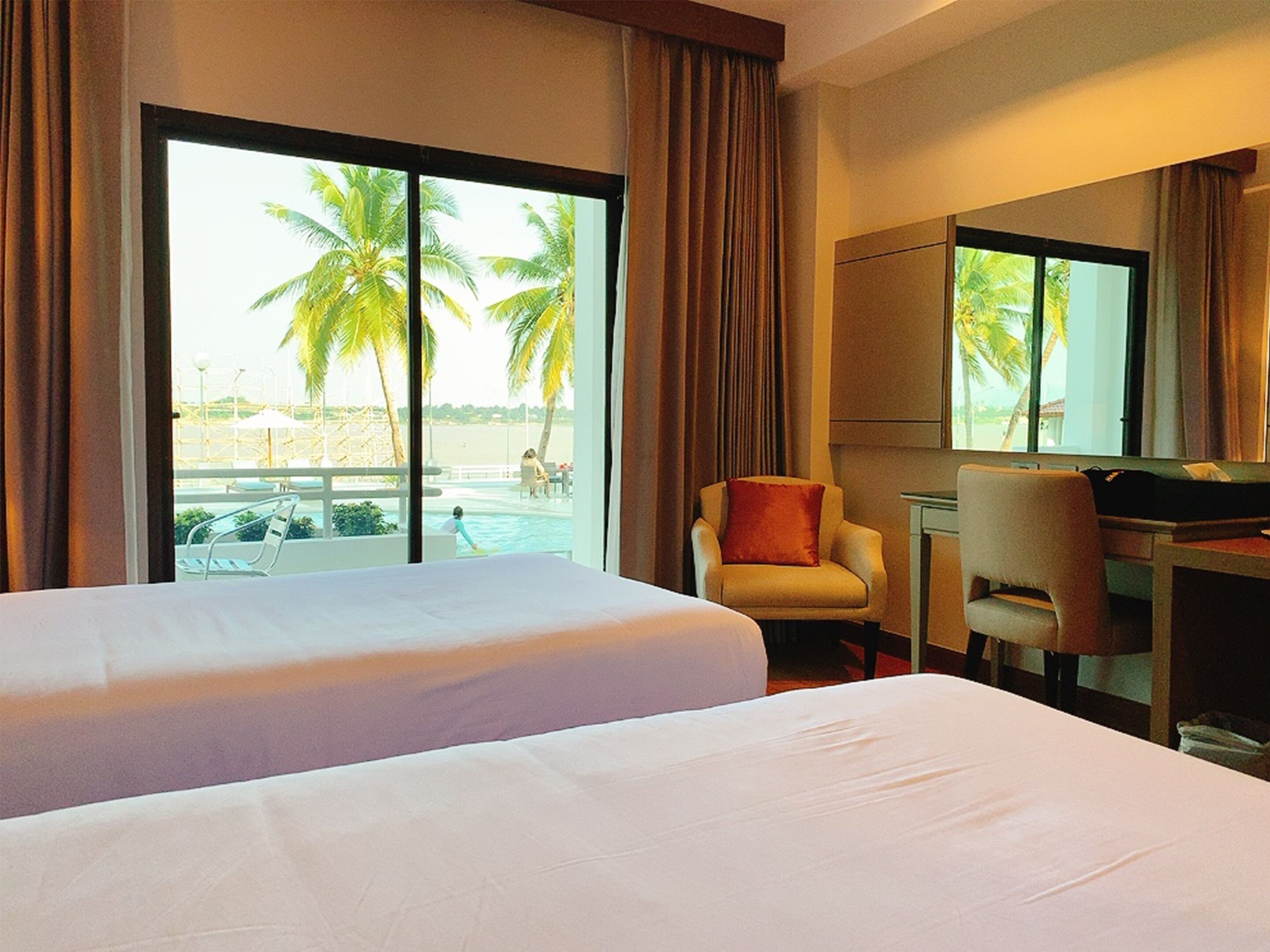 A hotel room with two beds, a desk, a chair, and a large window with a view of palm trees and a pool.