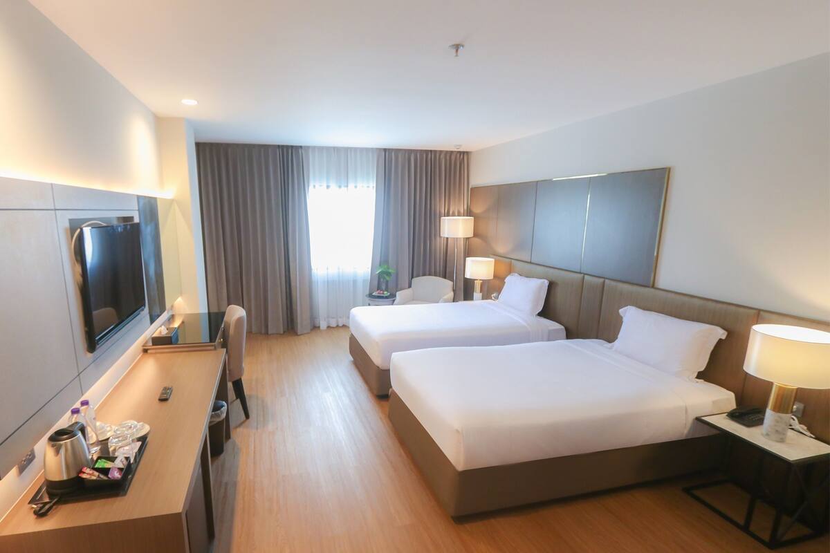 Habitación de hotel con dos camas, un escritorio con televisor, una silla y una mesita de noche con lámpara.