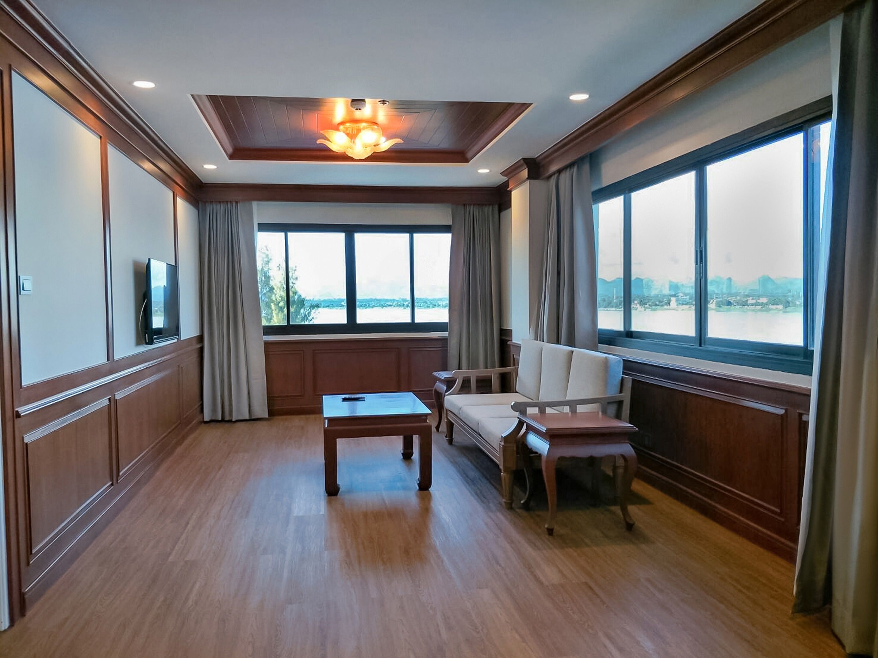 A spacious room with a large window offering a view of a body of water and distant buildings. There is a sofa, a small table, and a television mounted on the wall.