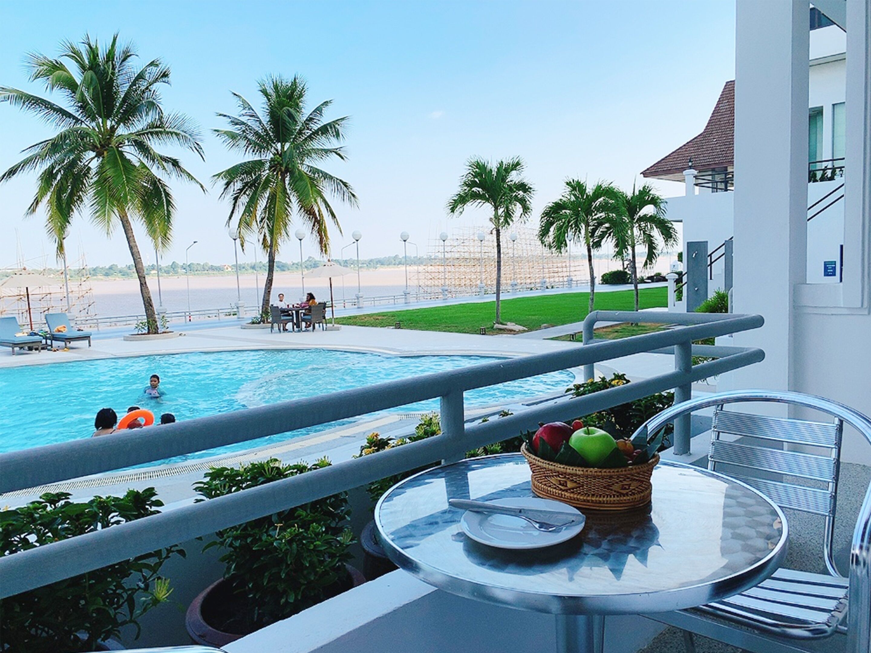 A poolside view with a dining area, palm trees, and a beach in the background.