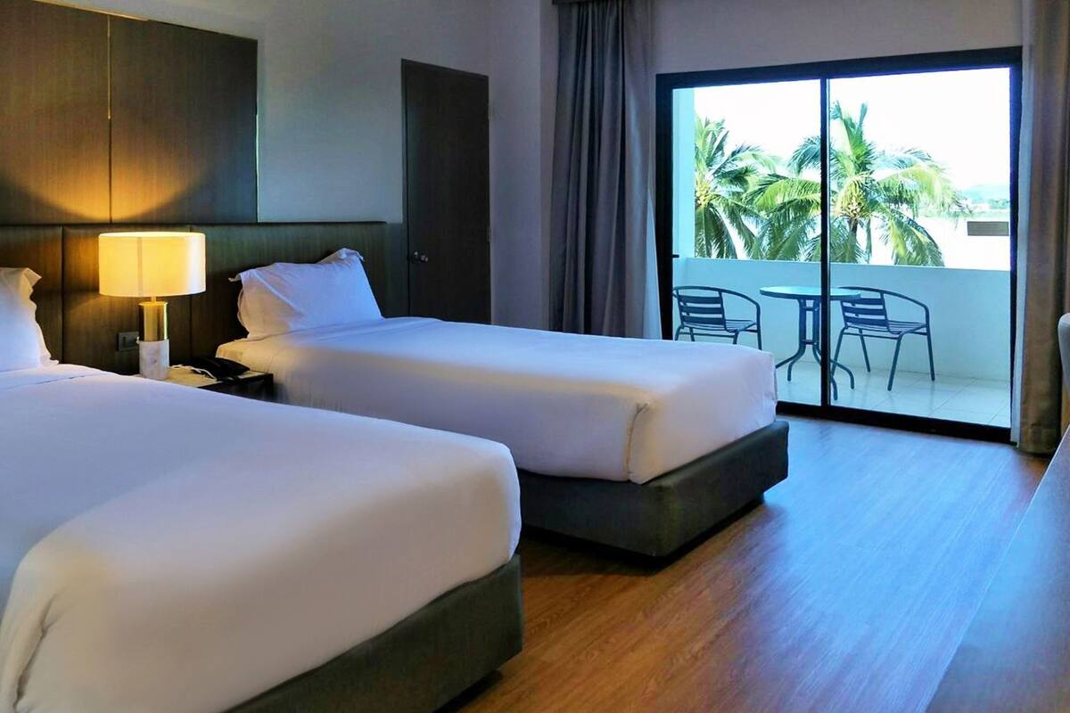 Habitación de hotel con dos camas, una mesita de noche, una lámpara, una puerta corrediza de vidrio a un balcón con sillas y vistas a palmeras.