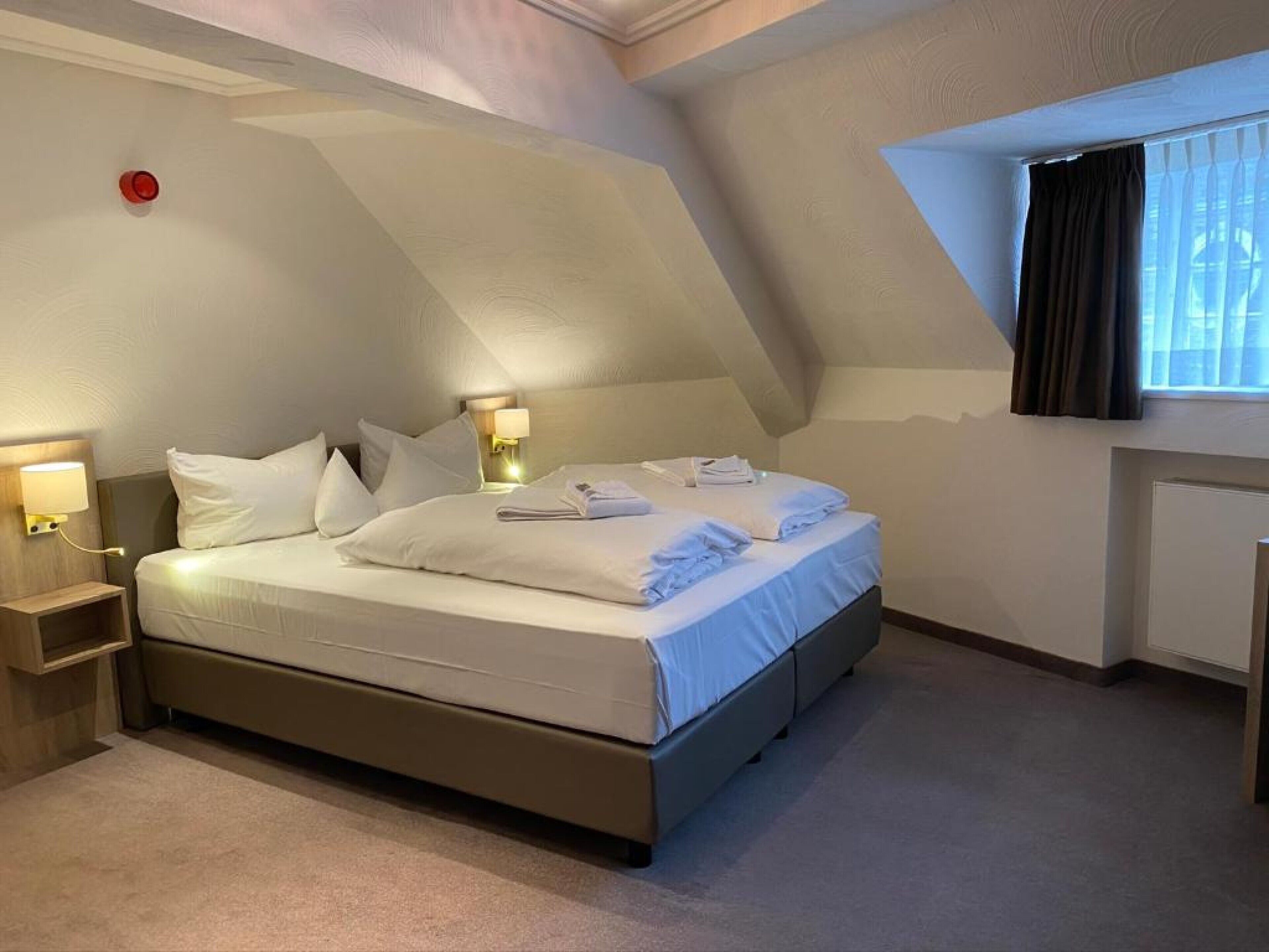 Een net opgemaakt bed in een kamer met een schuine plafond, een bedlampje en een raam met gordijnen.