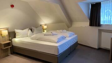 Een net opgemaakt bed in een kamer met een schuine plafond, een bedlampje en een raam met gordijnen.