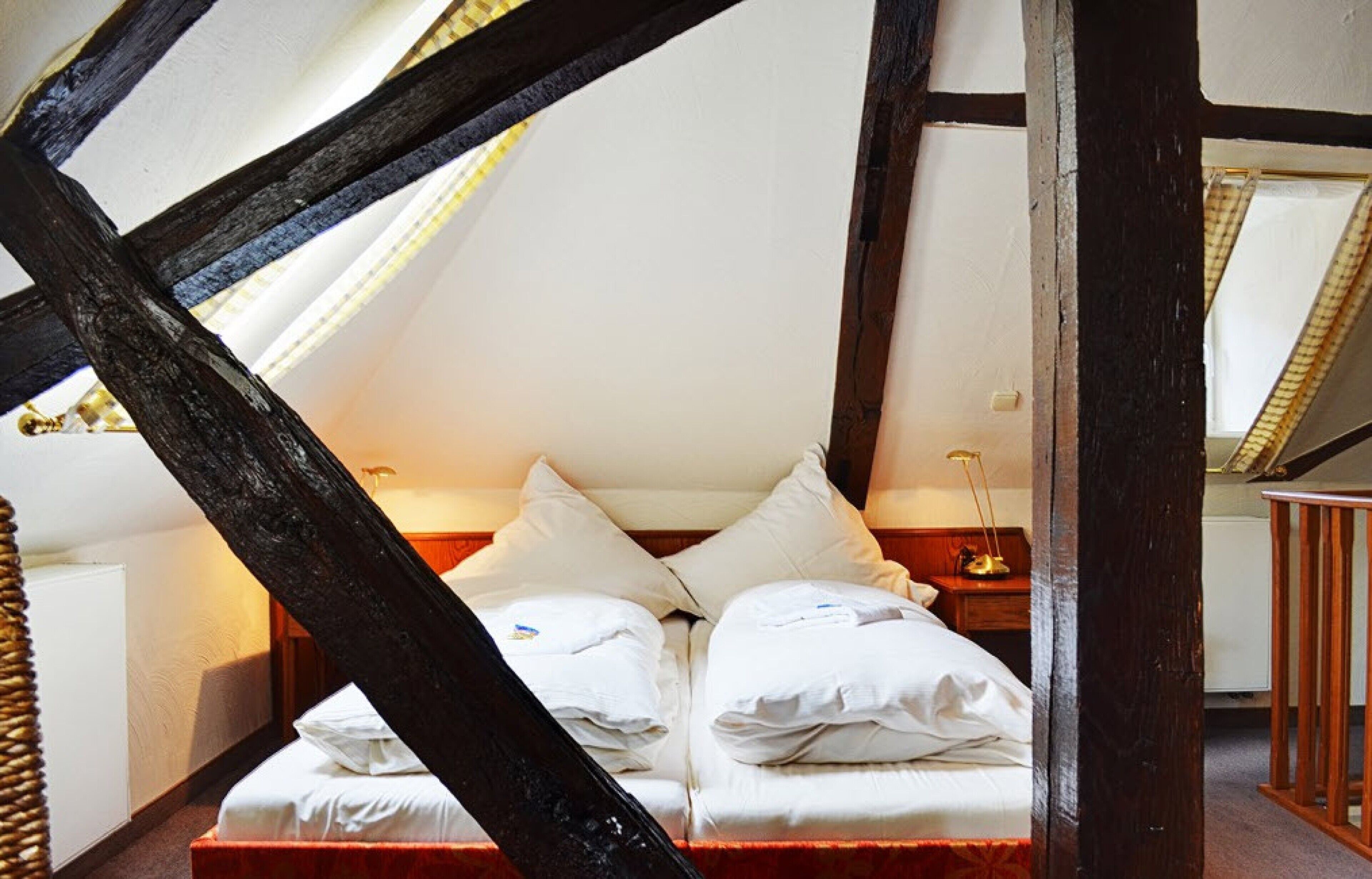 A bedroom with a high ceiling and exposed wooden beams, a bed with white linens, and a bedside table with a lamp.
