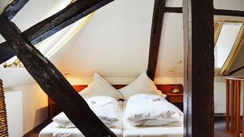 Quarto com teto alto e vigas de madeira aparentes, cama com roupa de cama branca e mesa de cabeceira com abajur.