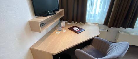 A hotel room with a wooden desk, a television, a chair, a bottle, and a book.