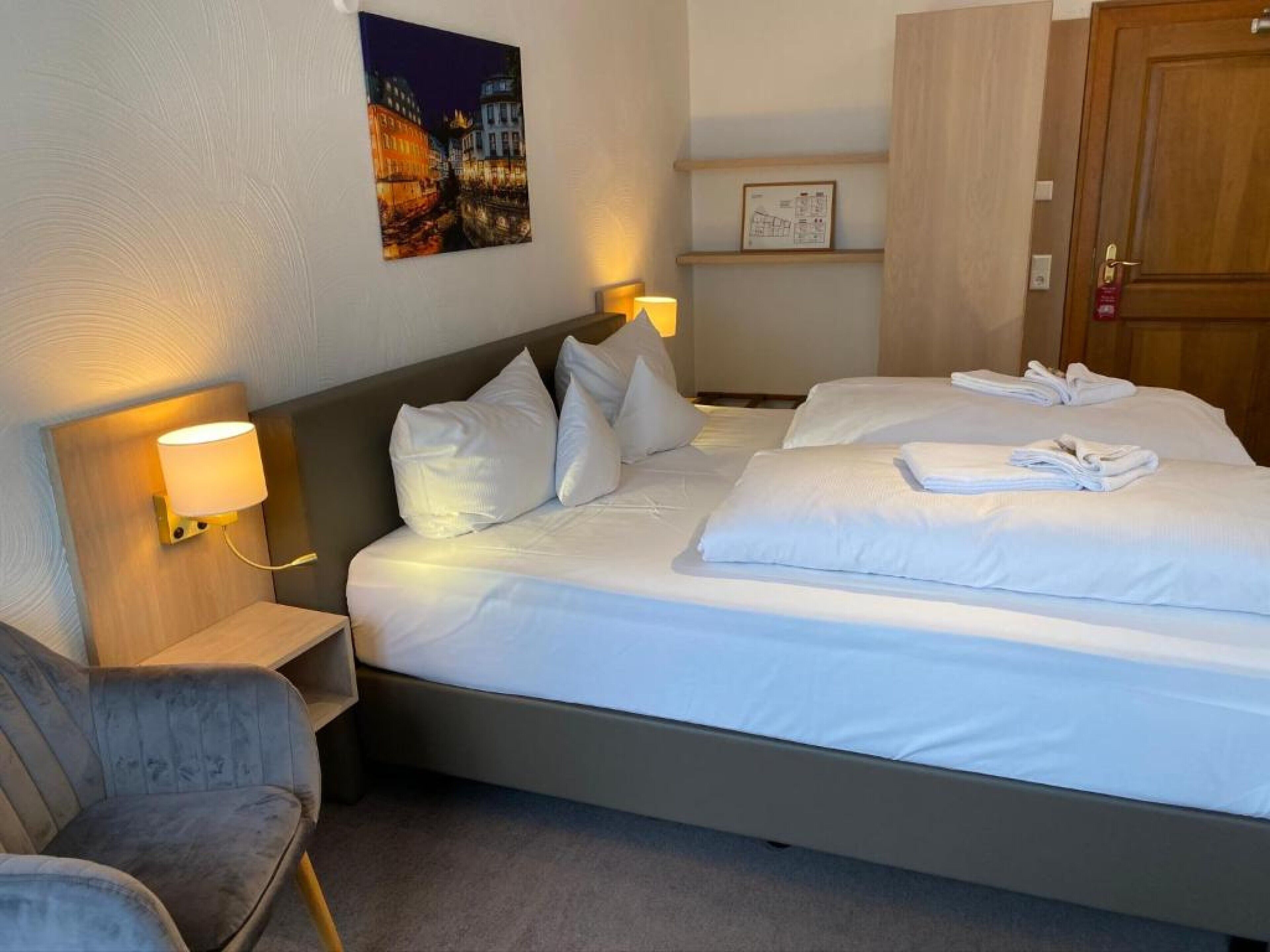 A hotel room with two beds, a chair, a nightstand with a lamp, a framed map on the wall, and a wooden door.
