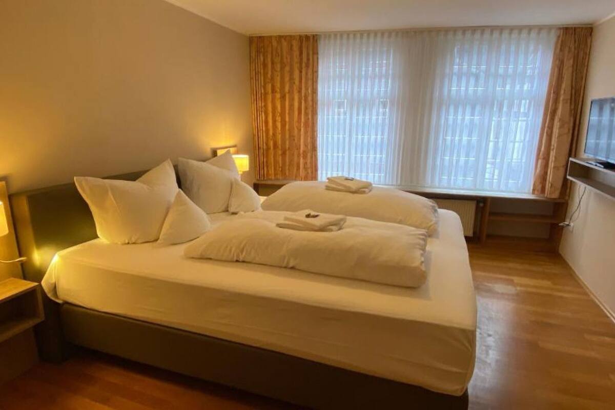 Ein Hotelzimmer mit einem großen Bett, zwei Nachttischen mit Lampen, einem flachbildfernseher an der Wand und einem Fenster mit Vorhängen.