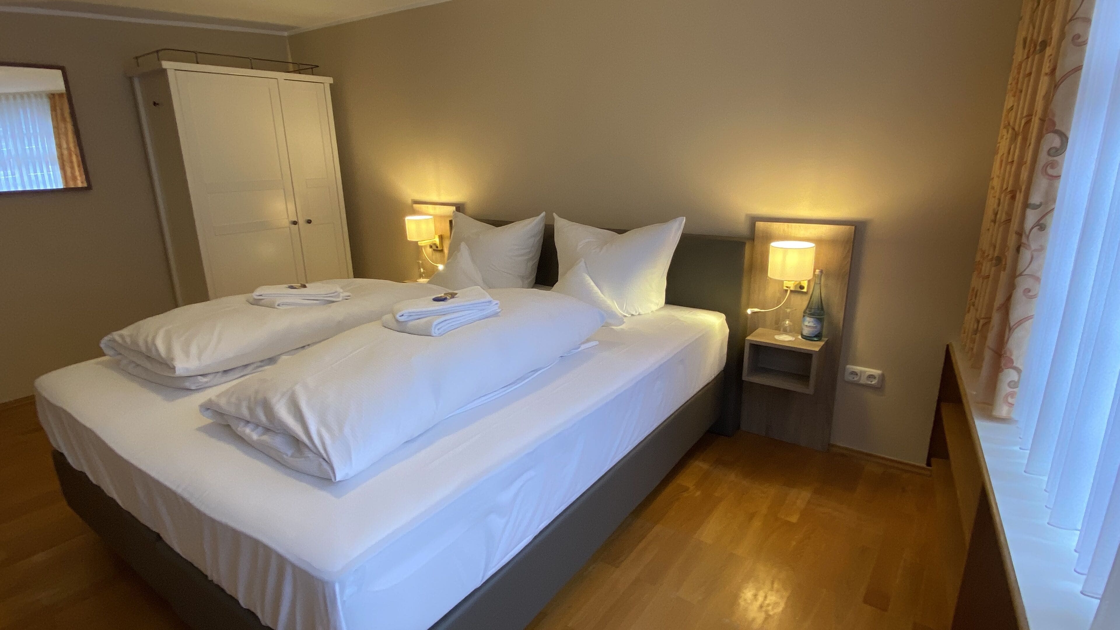 Una cama doble bien tendida con sábanas y toallas blancas, dos mesitas de noche con lámparas, y un armario al fondo.