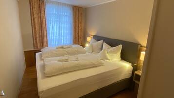 A neatly made bed with white linens and pillows, a bedside lamp, and a window with curtains.