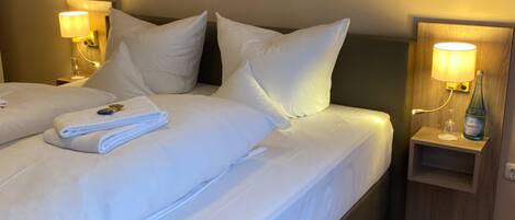Ein Hotelzimmer mit zwei Betten, jeweils mit weißer Bettwäsche und Kissen, einem kleinen Nachttisch mit einer Wasserflasche sowie Wandlampen.