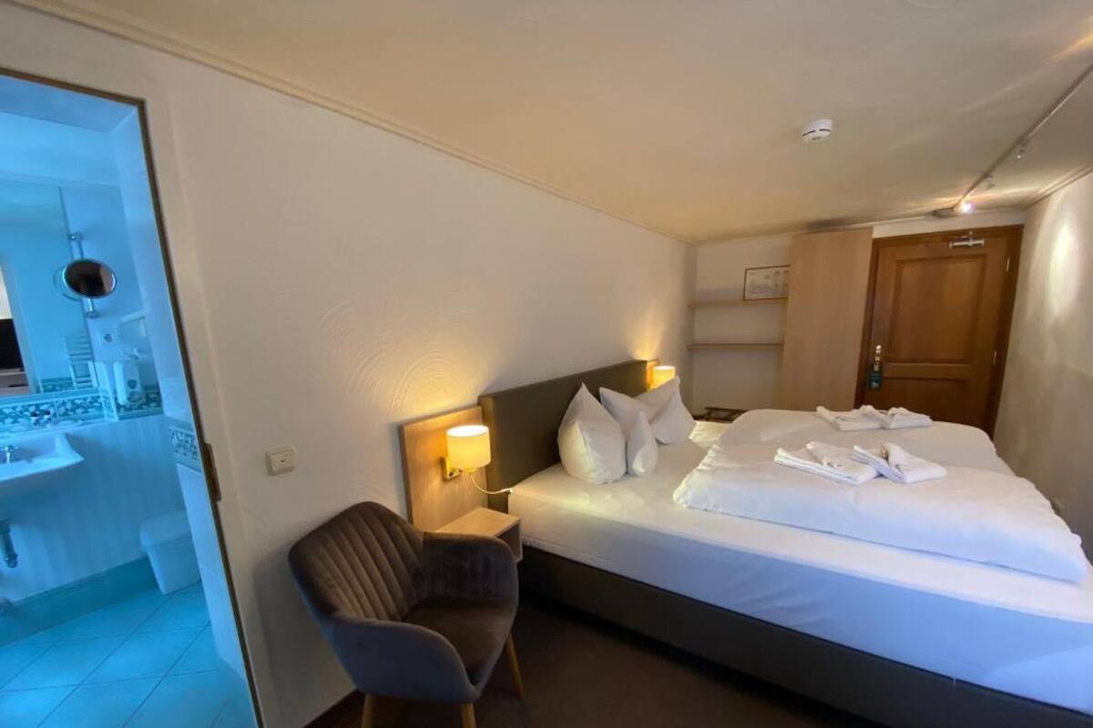 Ein Hotelzimmer mit Bett, Nachttisch, Lampe, Stuhl und einem durch eine offene Tür einsehbaren Badezimmer.