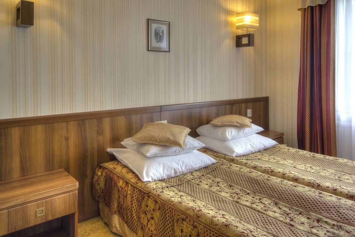Une chambre d’hôtel avec deux lits, des têtes de lit en bois, une table de chevet, un tiroir et un luminaire fixé au mur.