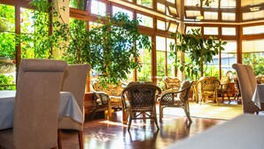 Een restaurant met rieten stoelen en tafels, grote ramen en binnenshuis planten.