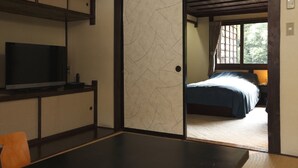 A traditional Japanese-style room with tatami flooring, a low table, a television, and a bed with a blue bedspread. - Hazuki (Shinshiro)