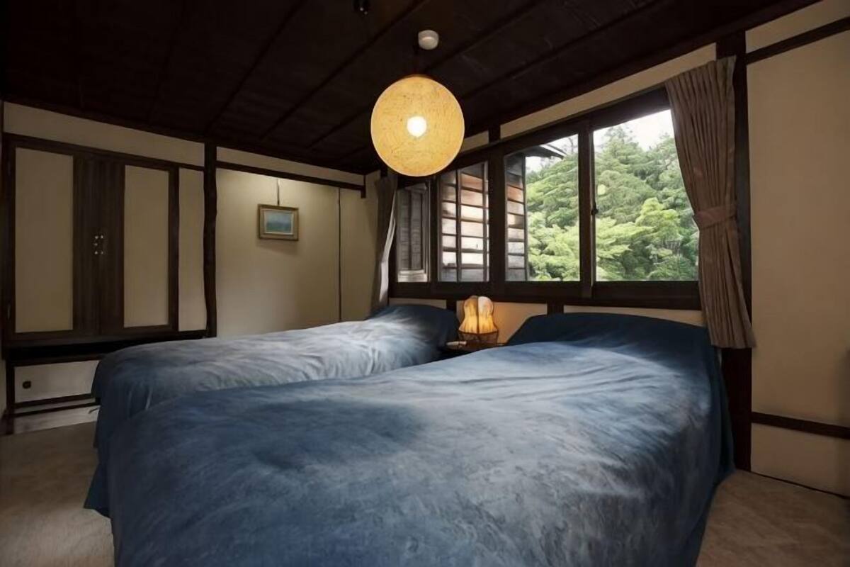 A traditional Japanese-style room with tatami flooring, a low ceiling, and a large window with a view of greenery. There are two beds with blue bedding and a hanging light fixture.