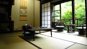A traditional Japanese room with tatami flooring, a low table, a chair, and a view of greenery through large windows. - Hazuki (Shinshiro)