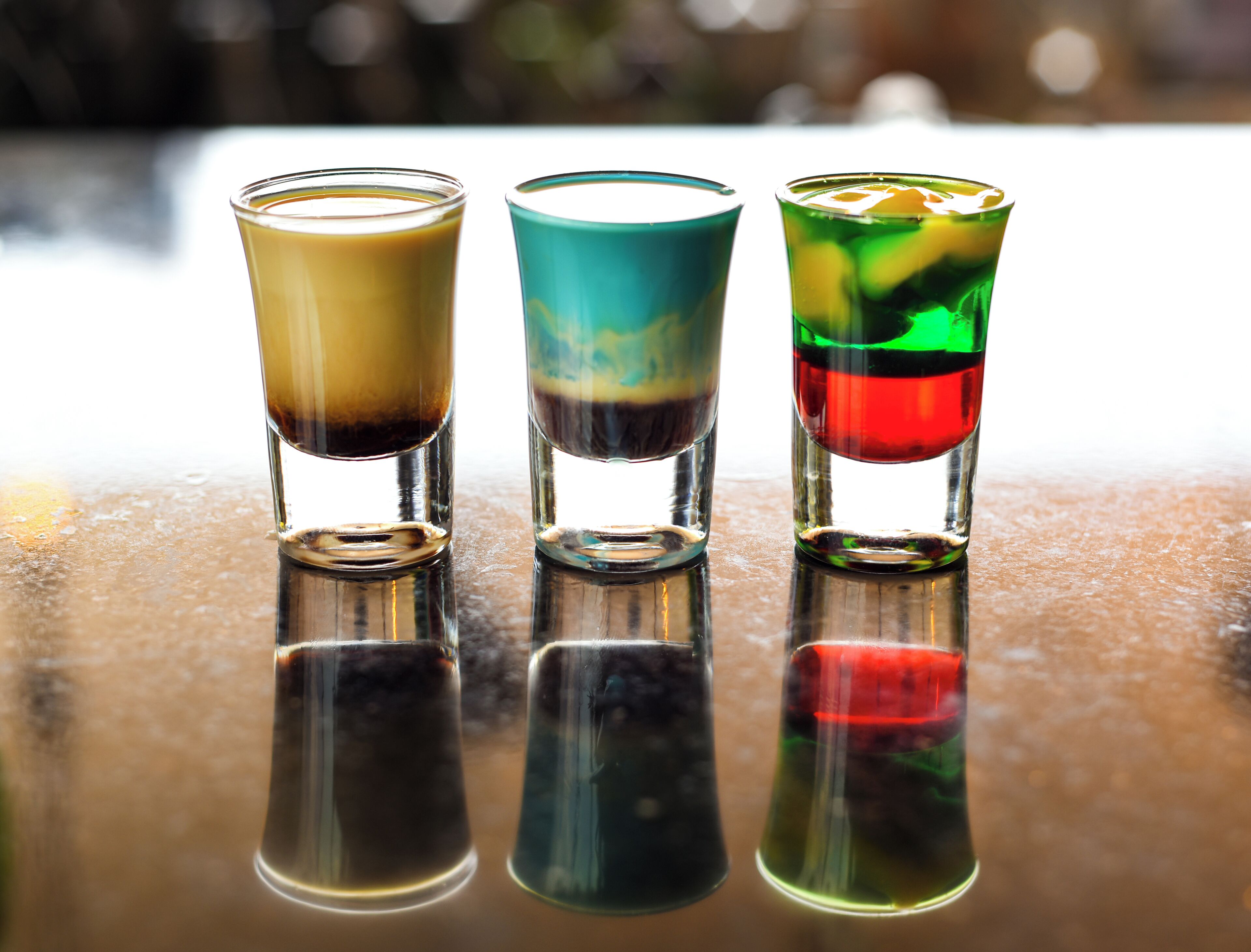 Three shot glasses with layered drinks on a reflective surface.