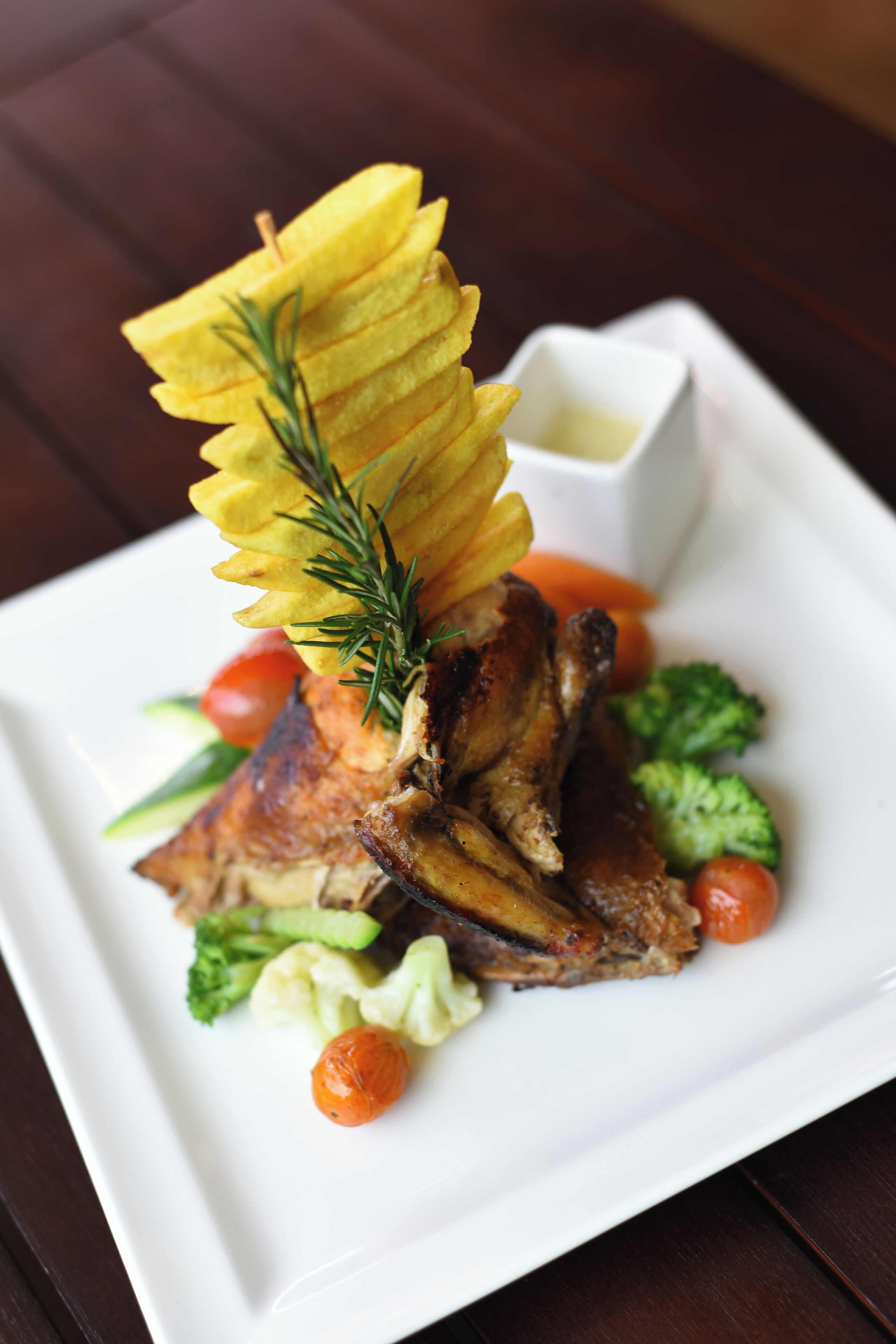 A plate of food with a roasted meat centerpiece, garnished with a sprig of rosemary, surrounded by vegetables and a small bowl of sauce.