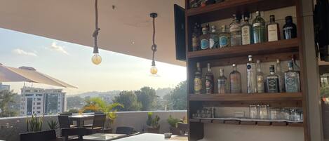 A rooftop bar with a variety of liquor bottles on shelves, hanging light bulbs, and outdoor seating with tables and chairs.