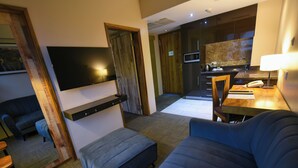 A modern hotel room with a flat-screen TV mounted on the wall, a sofa, a small table, and a kitchenette area. - Arcadia Suites (Kampala)
