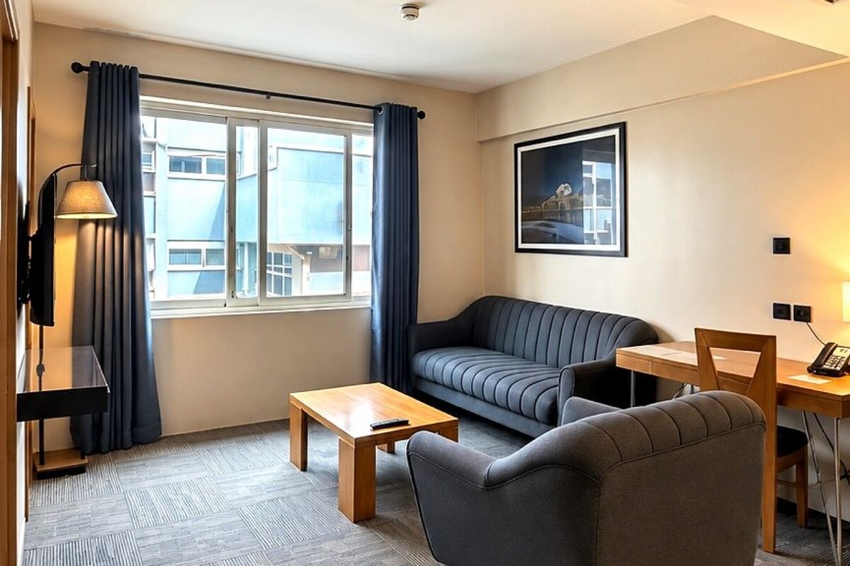 A modern hotel room with a sofa, a small table, a desk with a lamp, and a window with curtains.