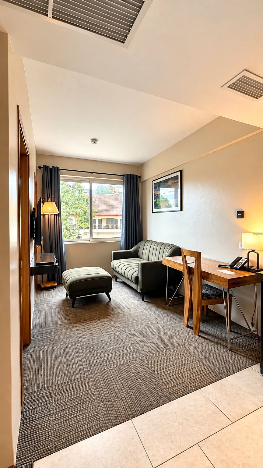 A hotel room with a sofa, a wooden table, a lamp, a desk, a telephone, and a window with curtains.