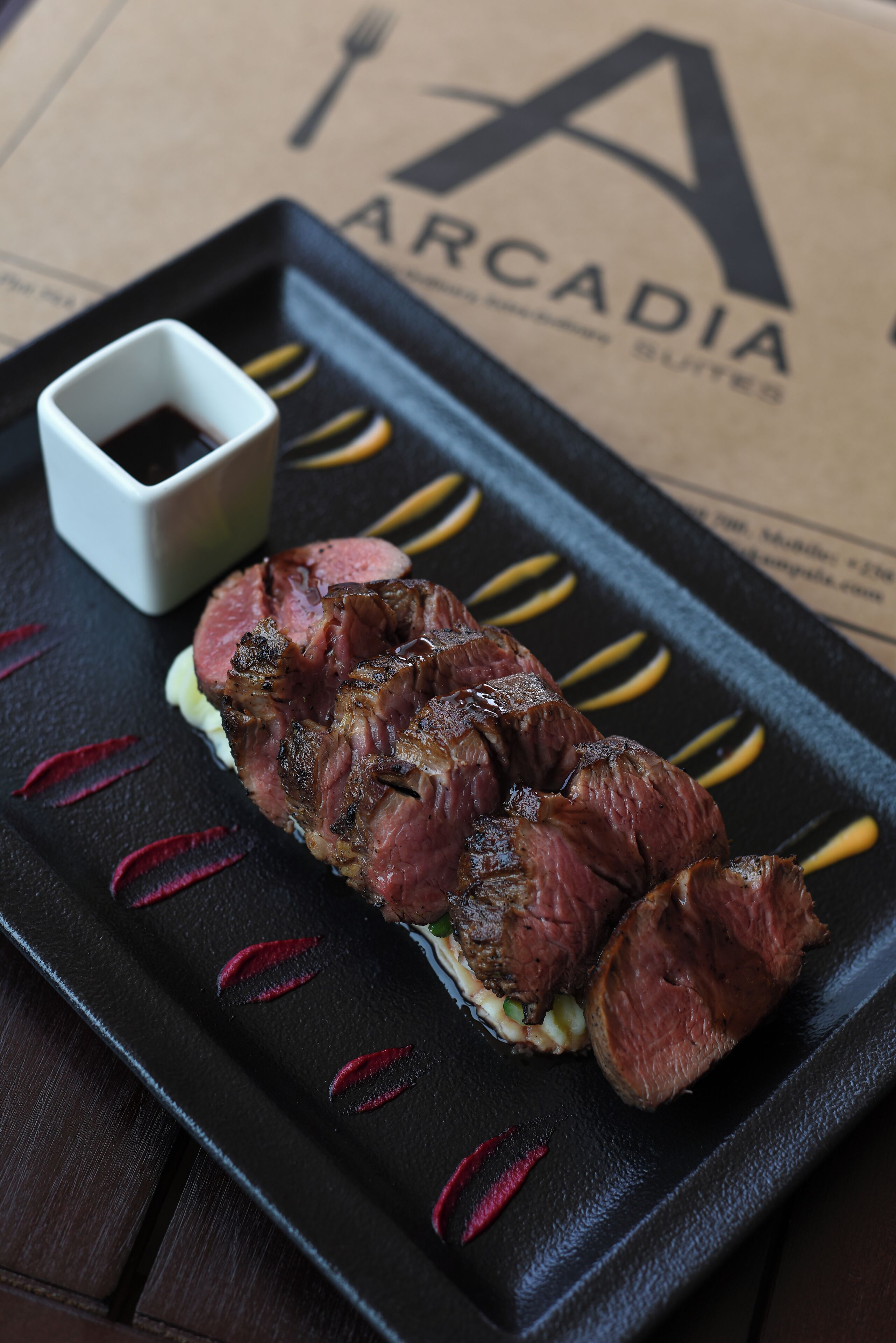 A steak with a side of sauce on a black plate, placed on a table with a paper featuring the word "ARCADIA" in the background.