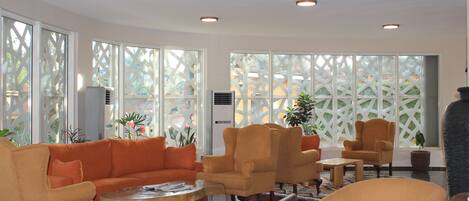 Eine Lobby mit orangefarbenen Sofas, Sesseln und einem Couchtisch. Große Fenster mit Gittermuster, und Zimmerpflanzen sind sichtbar.