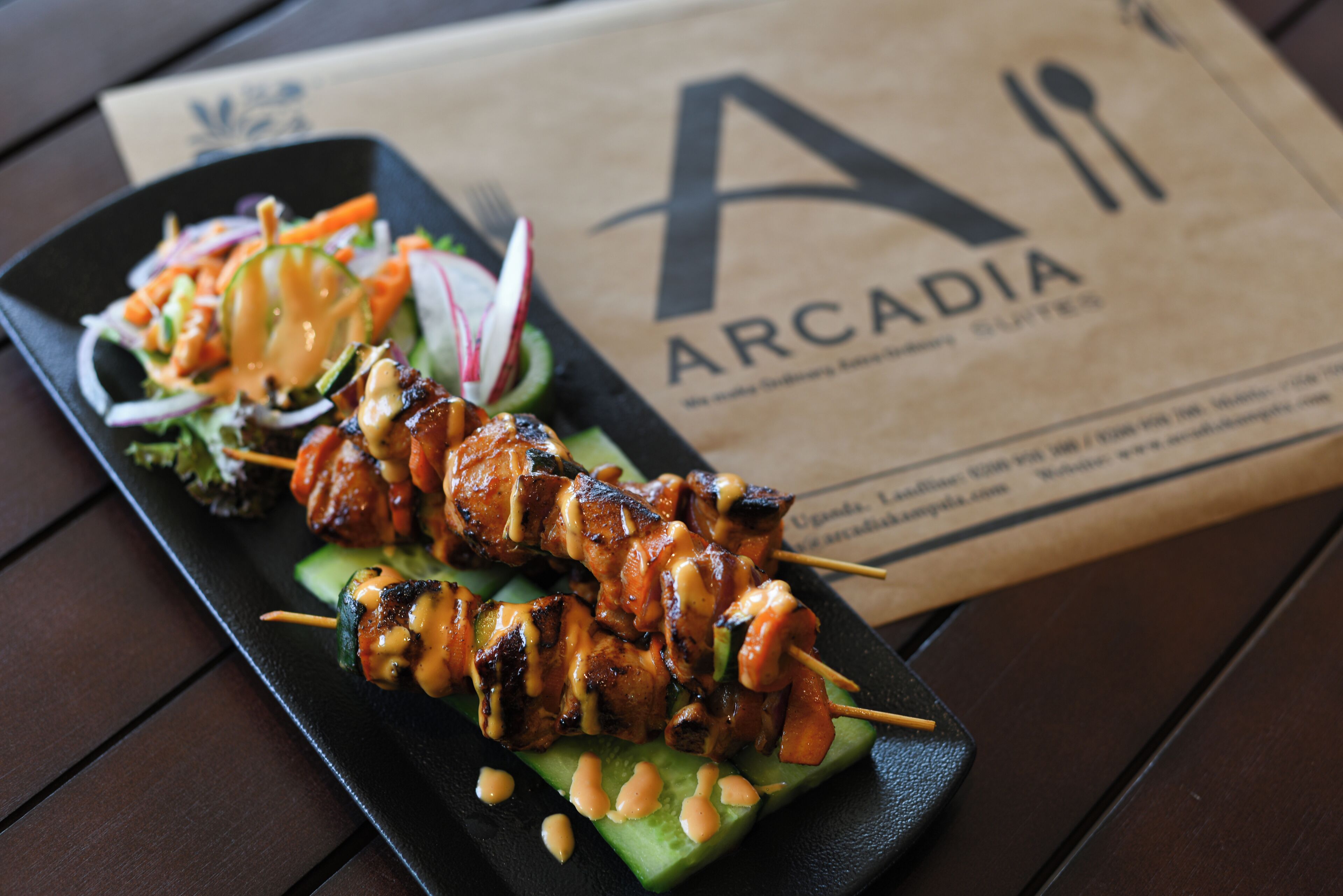 A plate of skewered meat with vegetables and a side salad, placed on a black plate with a brown paper bag labeled "ARCADIA" in the background.