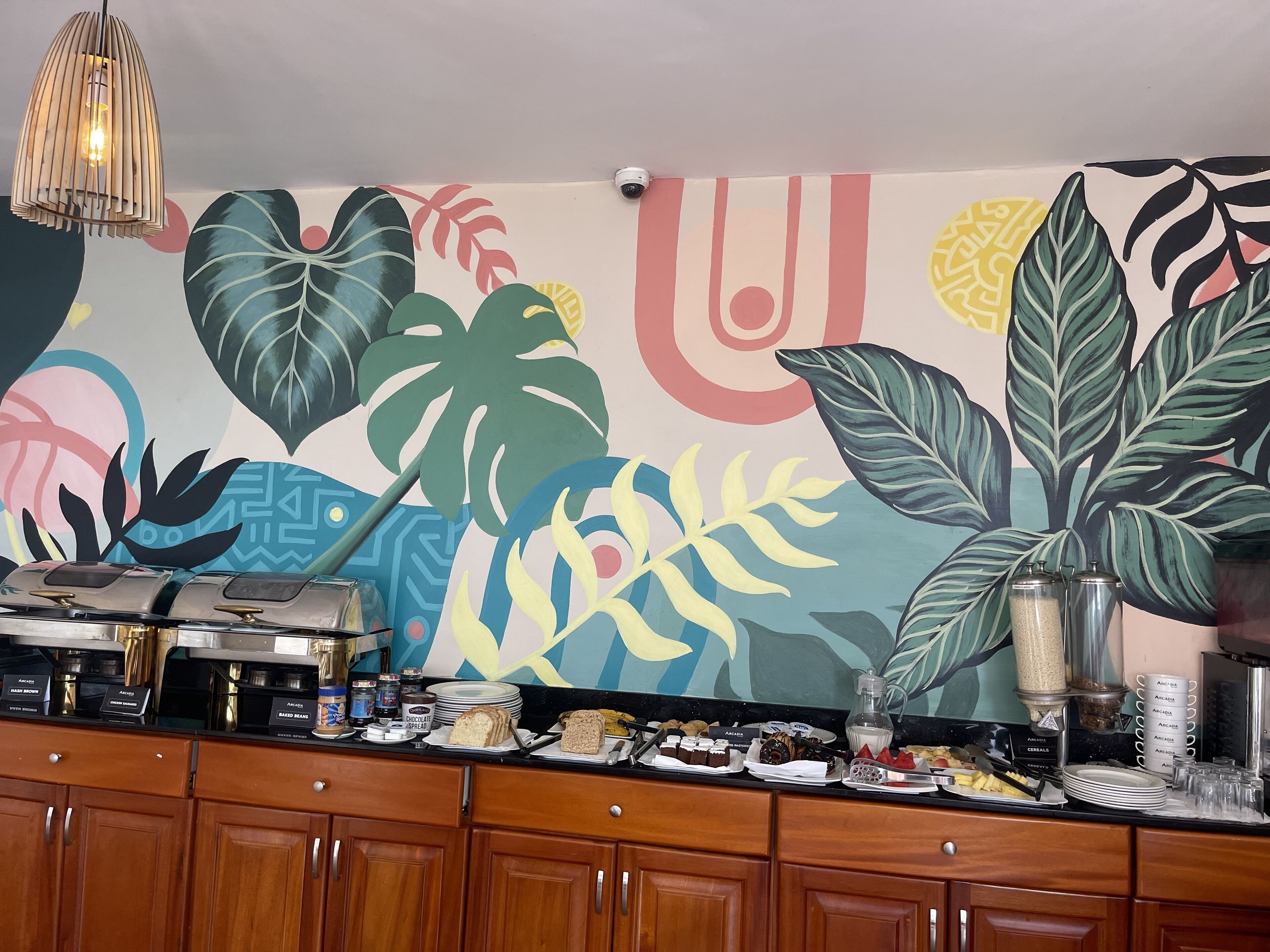 Zona de bufé con un mural tropical lleno de color en la pared, armarios de madera debajo y diferentes alimentos expuestos en la barra.